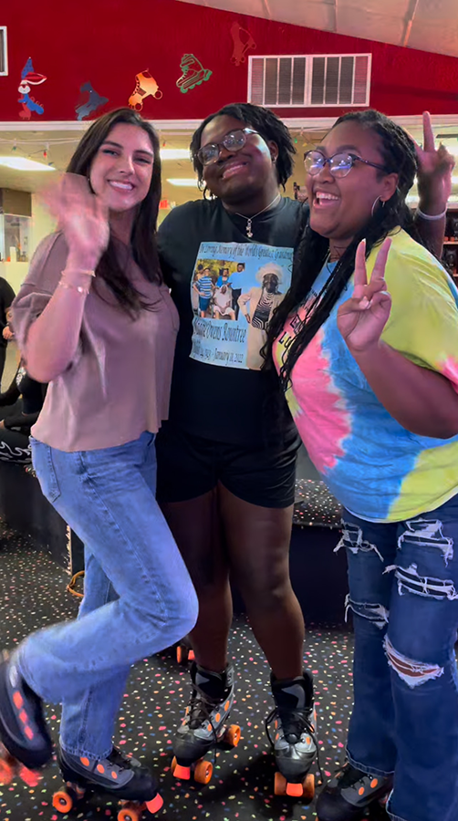 Three women pose for a photo while wearing roller skates inside a skating rink.